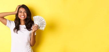 Güzel bir Afro-Amerikalı kız resmi, para kazanıyor, mutlu bir şekilde gülümsüyor gözleri kapalı, kendini şanslı hissediyor, sarı arka planda duruyor..