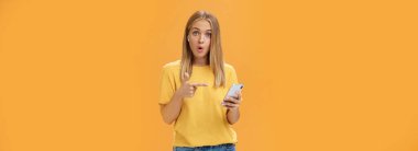 Akıllı telefonlara bağlanan kablosuz kulaklıklardan etkilenen adam ekranı işaret ederek sorgulanıyor ve şaşırtılan kıvrılmış dudaklar turuncu duvarın üzerinde kulaklık takan kameraya bakıyor.