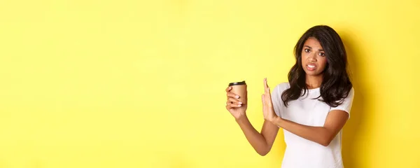 Image of african-american girl complaining about bad taste of coffee, showing reject sign and pulling away cup, standing over yellow background.