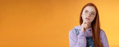 Düşünceli, genç, yaratıcı, zeki, kızıl saçlı kız sol üst köşede durup, gözlerini kısarak düşünmeyi, dokunmayı düşünmeyi, düşünmeyi, turuncu arka planda kalmayı önemli buluyor..