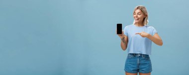 Sarışın, güzel Avrupalı bir kadının portresi. Şaşkınlıkla akıllı telefon ekranını gösterirken mavi arka planda yepyeni bir aleti tanıtıyor.