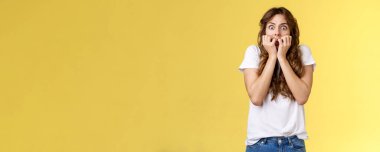 Şok olmuş, konuşamayan, korkmuş, kıvırcık saçlı, beyaz kadın nefes nefese, parmaklarını tutuyor, tırnak yiyor, kameraya bakıyor, korku dolu korku filmi sarı arka planda duruyor..