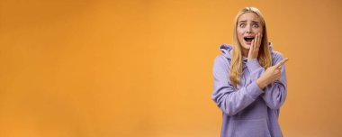 Şaşırtıcı derecede heyecanlı kapüşonlu genç sarışın kız avucunun içi gibi bağırıyor sağa doğru hayret verici bir şekilde haykırıyor ünlü süper star neşeli, pozitif duyguları tutamıyor, turuncu arka plan.