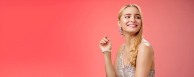 Zarif, göz kamaştırıcı, genç, zengin, sarışın bir kadın, şık, gümüş renkli bir aksesuarla yardım partisine katılıyor sağa dönüyor, gülümsüyor, tanıdık birini selamlıyor, neşeli, kırmızı arka planda sırıtıyor..