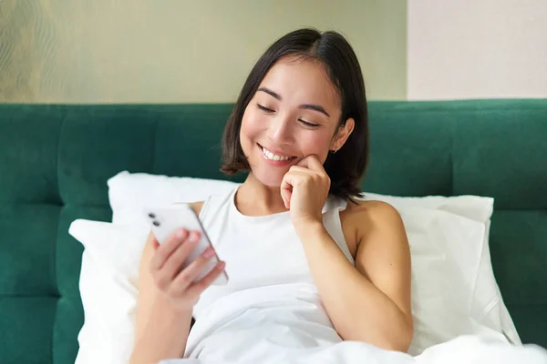 Beautiful korean woman reading mobile phone, messaging or watching video while relaxing in her bedroom, holding smartphone.
