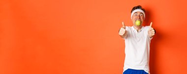 Orta yaşlı, spor kıyafeti giymiş, elmayı ağzında tutan, parmaklarını kaldırıp turuncu arka planda duran komik bir adam resmi..