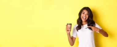 Afro-Amerikalı neşeli bir kadın tasviri, kafeden iyi kahve öneriyor, fincanı işaret ediyor, sarı arka planda duruyor..