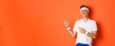 Spor yapan aktif ve sağlıklı bir adamın portresi, aktif giysi giyen, gülümseyen ve parmakla işaret eden, turuncu arka planda duran..