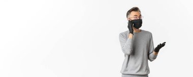 Covid-19 kavramı, sosyal mesafeler ve yaşam tarzı. Medikal maskeli ve eldivenli yakışıklı orta yaşlı bir adamın görüntüsü, cep telefonuyla konuşuyor, beyaz arka planda duruyor..