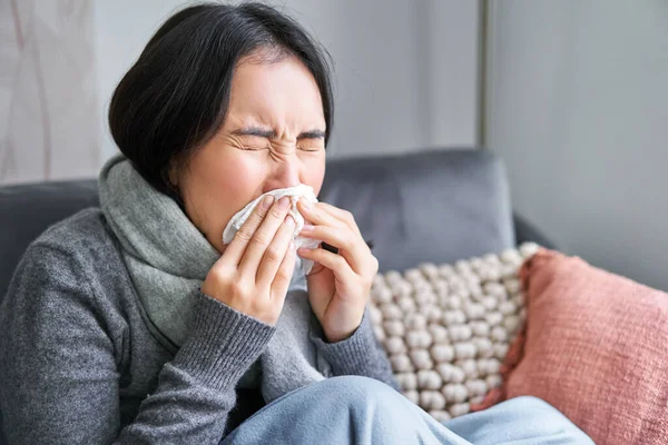 Close up of young korean woman staying at home with cold, sneezing in napking, has runny nose, concept of illness, health and influenza.