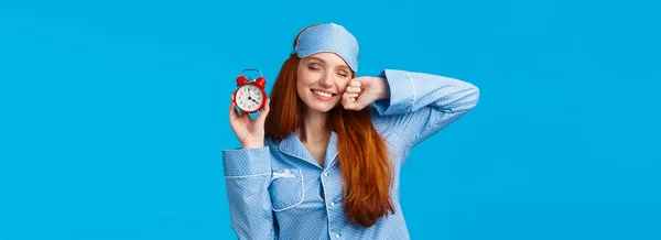 Girl slept well, wake up energized, sleepy stretching with closed eyes and lovely smile, holding red cute clock, set up alarm to be work in time, wearing pyjama and sleep mask, blue background.