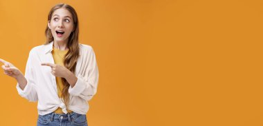 Yaşam tarzı. Tişörtünün üstünde dalgalı, açık renk saçlı, solu işaret eden, turuncu arka planda poz veren neşeli ve heyecanlı beyaz kız portresi..