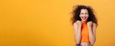 Mutlu ve zafer kazanan kıvırcık saçlı kadın. Kesilmiş üst yumruklarıyla evet hareketi yaparak gözlerini kapatıyor ve gülümsüyor. Harika haberler alıyor, turuncu arka planda ödül kazanıyor. Boşluğu kopyala