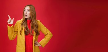 Yaşam tarzı. Empatik bir portre, şaşırmış ve şaşırmış sarı montlu kızıl saçlı kadın ağzı açık, şaşkın ve nutku tutulmuştu. Sol üst köşeyi işaret ediyor ve endişeli ve kırmızı duvarın üzerinden titriyordu..