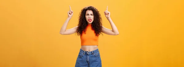 Indoor shot of displeased female client with curly stylish hairstyle. red lipstick in cool cropped top pointing up demanding explanation with confused and dissatisfied expression over orange