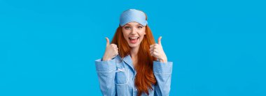 Belden yukarı portre zevkli, tamamen tatmin olmuş, uyku kıyafetleri, gece giysisi ve uyku maskesi takan sevimli kızıl kız, onaylanmış baş parmaklar gösteriyor, jest gibi, ya da aynı fikirde, mavi arka planda dikilen.