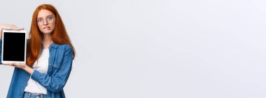 Ayartılmaya karşı koyamazsın, şimdi arabaya ekle. Çekici ve ilginç bakışlı, gözlüklü, heyecanlı kızıl saçlı, ısıran dudaklı, internetten bir şeyler alan, dijital tablet ekranı gösteren, beyaz arkaplan.