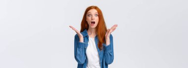Şaşırmış, dilsiz, kızıl saçlı bir kadın mükemmel haberler öğreniyor, hayallerin gerçekleştiğine inanamıyor, kameraya bakıyor, çene sallıyor, nefes nefese kalıyor ve el kaldırıyor şaşkınlıkla, beyaz arka plan.