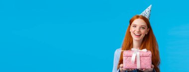 Hediye zamanı. Neşeli kızıl saçlı genç, kolejli kız doğum gününü kutluyor, elinde sevimli pembe bir hediye tutuyor ve doğum günü şapkası takıyor, neşeyle gülümsüyor mavi arka planda..