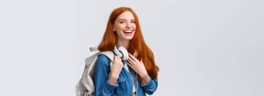 Eğitim, öğrenci hayatı ve mutluluk konsepti. Neşeli, güzel, kızıl saçlı, beyaz kız, sırt çantalı ve kulaklıklı üniversite öğrencisi kameraya gülümsüyor..