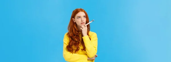 Troubled, hesitant and indecisive redhead, ginger girl with curly hair in yellow sweater, looking unsure upper left corner as pondering, solving exercise in mind, holding pen, searching inspiration.