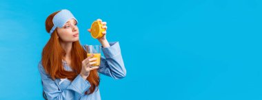 Uyku maskesi ve geceliği takmış, meyve yemek gibi umut dolu ve hevesli bir ifadeyle kahvaltılık meyve suyu, mavi arkaplan yapmak için cama portakal sıkıştıran aptal ve sevimli kızıl saçlı beyaz kız..