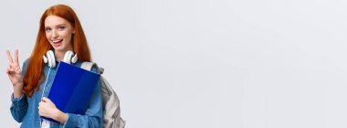 Sevimli, arkadaş canlısı üniversite öğrencisi, evrakları olan kızıl saçlı, okul için evrakları olan, sırt çantası taşıyan ve boynuna kulaklık takan, resmi olmayan bir selamlaşma hareketi yapan, beyaz bir geçmişi olan bir kız..