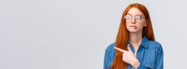 Gözlüklü, zeki ve sevimli kızıl saçlı genç, dudak ısıran kız öğrenci merakla bakıyor ve sol tarafı işaret ediyor, düşünüyor, karar veriyor, seçim yapıyor, beyaz arka planda duruyor..
