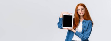 Ayartılmaya karşı koyamazsın, şimdi arabaya ekle. Çekici ve ilginç bakışlı, gözlüklü, heyecanlı kızıl saçlı, ısıran dudaklı, internetten bir şeyler alan, dijital tablet ekranı gösteren, beyaz arkaplan.