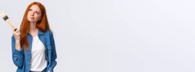 Hayalperest ve kararsız kızıl saçlı kadın kot gömlekli, sırıtan ve gözlerini kısan düşünceli bir şekilde bir şeyler hayal eden, elinde fırça tutan, boyayı hangi renge boyayacağını düşünen..