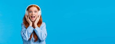 Heyecanlı ve eğlenceli beyaz saçlı, pijama giymiş çekici bir kadın, hayranlık ve şaşkınlıktan bağırıyor, yeni bir şarkı duyuyor, kulaklık takıyor, müzikten hoşlanıyor, mavi arka planda duruyor..