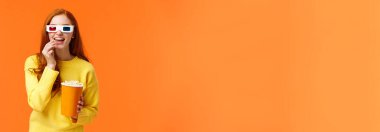 İlgili ve eğlenceli, sarı kazaklı güler yüzlü kızıl saçlı kadın, sinemada film izliyor, 3 boyutlu gözlük takıyor, patlamış mısır yiyor ve eğlenceli ve heyecanlı bir ifadeyle meraklı meraklı bakıyor..