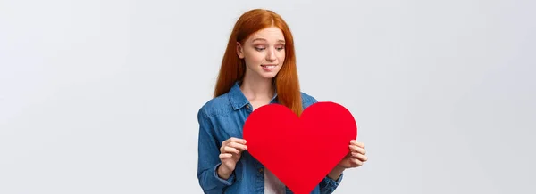 Indecisive cute and timid, shy redhead girl wants confess in love, show sympathy unsure give present on valentines day or not, holding big red heart, biting lip worried, white background.