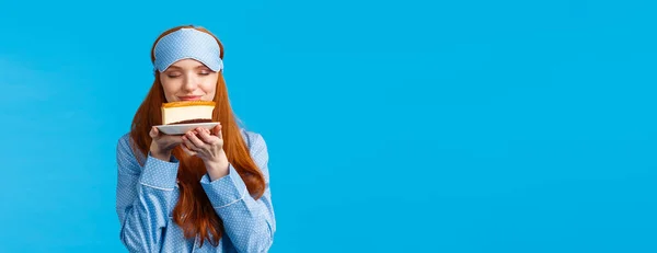 Food, eating and desserts concept. Cheerful pretty redhead woman in nightwear, sleep mask, close eyes smelling tasty slice cake want eat sweets but on diet, standing blue background.