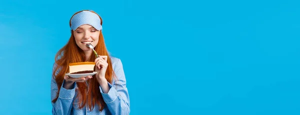 Girl feeling satisfaction eating sweet delicious dessert in morning. Attractive cute woman with red hair in nightwear and sleep mask, close eyes biting tasty cake, holding spoon close eyes.