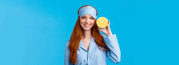Start your morning right. Diet, healthy lifestyle and beauty concept. Cheerful glamour redhead woman in nightwear, holding slice orange near face, smiling, wearing sleep mask and pyjama.
