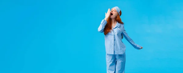What wonderful morning. Full-length vertical studio shot yawning cute caucasian redhead girl in sleep mask and pyjama, stretching and cover opened mouth with closed eyes, blue background.