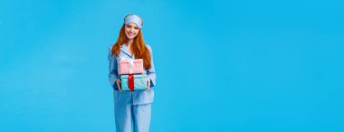 Neşeli ve güzel genç mutlu kızıl saçlı kadın Noel arifesinde uyanıp kış tatillerini kutluyor, pijama giyiyor ve uyku maskesi takıyor, hediyelerle dolu kutular tutuyor, hediyeler veriyor..