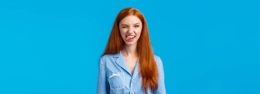 Şımarık ve heyecanlı, gece elbisesi içinde güzel saçlı bir kadın, neşeyle göz kırpan ve gülümseyen bir kamera, harika hissediyor, pozitif duygularını ifade ediyor, mavi arka planda duruyor..