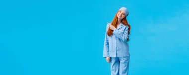 Dikey tam boy stüdyo, neşeli ve aptal, gece giysisi ve uyku maskeli hassas kadın, başını eğ ve sevimli bir gülümseme kamerasıyla bakıyor, sol üst köşeyi işaret ediyor, mavi arka plan duruyor..