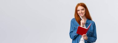 Yaratıcı, güzel, kızıl saçlı kız öğrenci, günlük kıyafetli genç, elinde kalem ve gülümseyen kamera, deftere notlar alıyor, günlük yazıyor, beyaz bir zemin hazırlıyor..