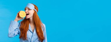 Lezzetli meyve sabah sağlıklı başlar. Belden yukarı portre çekici uykulu, uyku maskeli ve gecelikli güzel kızıl kız, portakal yiyen lezzetli, ayakta duran mavi arka plan.