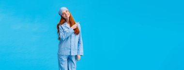 Dikey tam boy stüdyo, neşeli ve aptal, gece giysisi ve uyku maskeli hassas kadın, başını eğ ve sevimli bir gülümseme kamerasıyla bakıyor, sol üst köşeyi işaret ediyor, mavi arka plan duruyor..