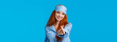 Hoş ve kadınsı, kızıl uzun saçlı güzel bir kız, uyku maskesi takmış ve pijama giymiş, narin gülümseyen ve başı eğilmiş, Kore kalbi ya da aşk jesti gösteren, mavi arka planda neşeli duran.