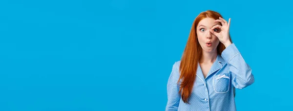 Wow amazing. Impressed and surprised curious redhead woman staring at something intriguing, looking through ok gesture folding lips and popping eyes startled, standing in pyjama blue background.