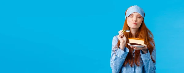 Delicious food, lifestyle and people concept. Delighted and happy cute redhead female in pyjama and sleep mask, close eyes and licking lips as eating tasty cake, holding spoon smiling.