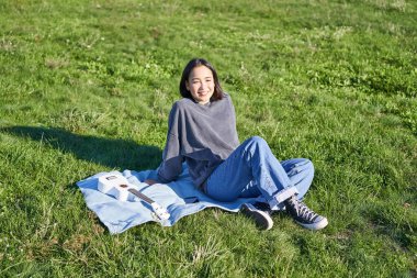 Parkta battaniyeye oturmuş gülümseyen tatlı kız, dışarıda ukulele oynuyor, güneşli havanın tadını çıkarıyor, kazak ve kot pantolon giyiyor..