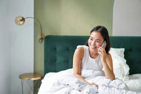 Smiling korean girl talks on mobile phone and lying in bed. Cute woman answers telephone call, holds smartphone, relaxing in her bed.