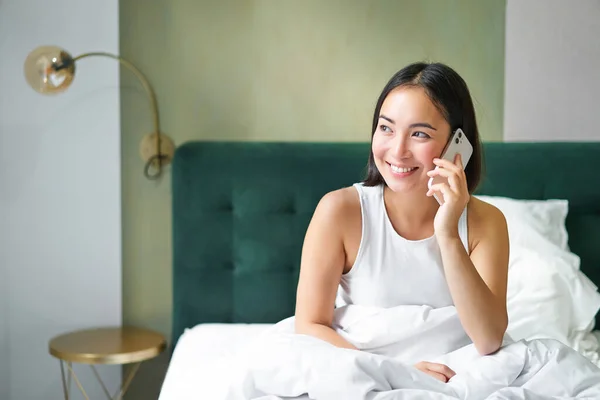 Smiling korean girl in bed, talks on mobile phone, making a phone call, lazy morning as asian woman orders delivery via smartphone.