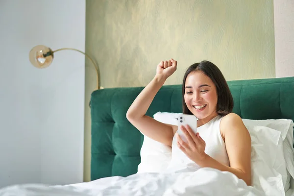 Enthusiastic asian girl reading mobile phone news, raising hand and triumphing in her bed, cheering while lying on pillow in bedroom, positive news.